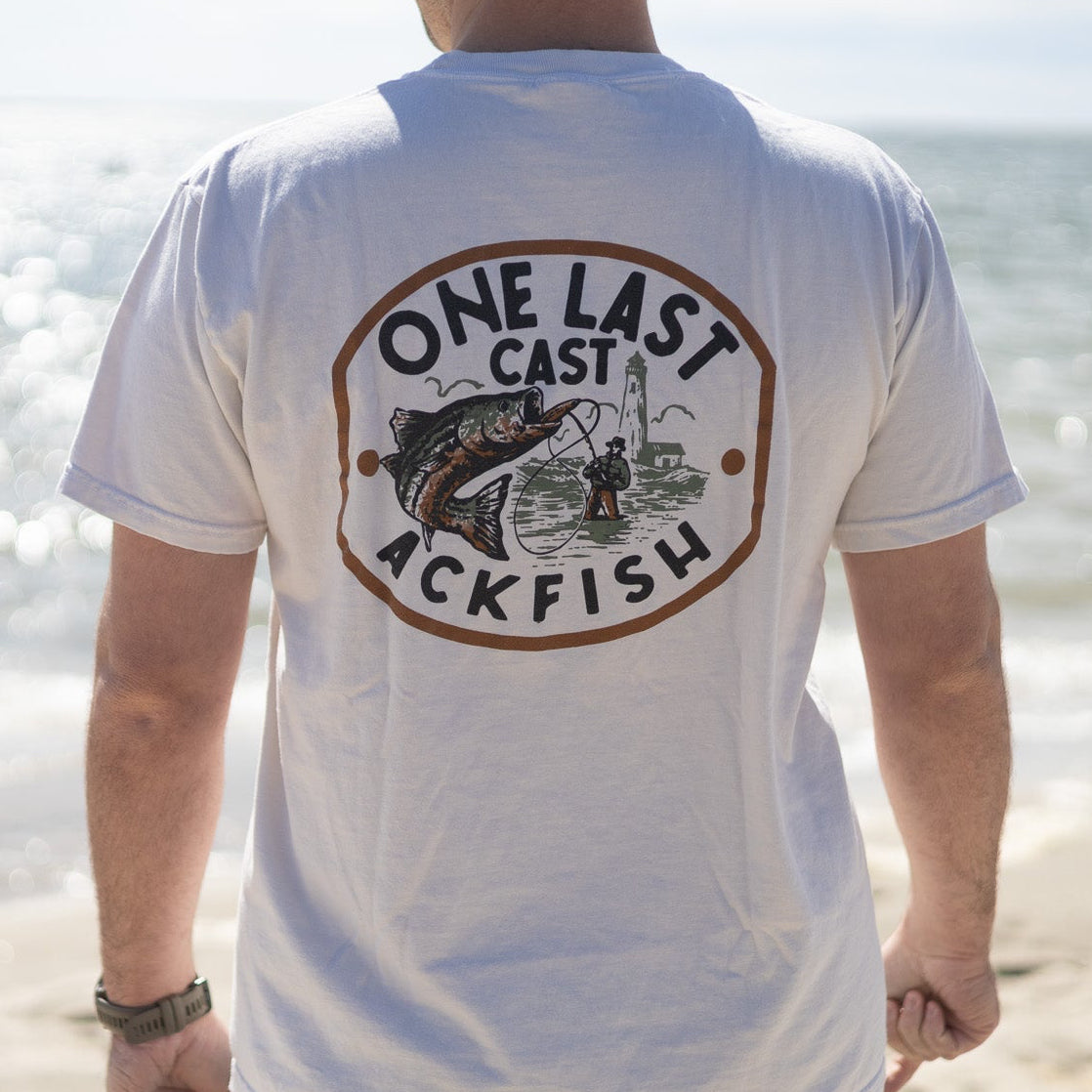 A man standing on a beach with his back to the camera, wearing a white t-shirt with a large circular graphic on the back that reads 'ONE LAST CAST ACKFISH' with an illustration of a fish and fishing rod.