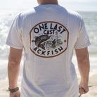 A man standing on a beach with his back to the camera, wearing a white t-shirt with a large circular graphic on the back that reads 'ONE LAST CAST ACKFISH' with an illustration of a fish and fishing rod.
